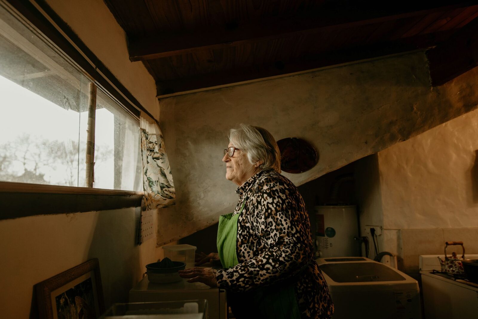 An elderly woman wearing glasses and a leopard print jacket looks out a window indoors.