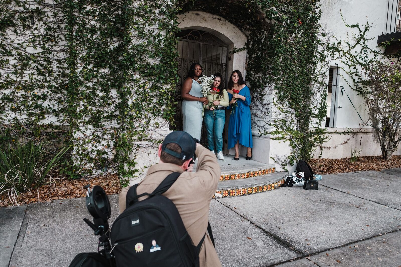 A photographer captures three women with floral bouquets on a vine-covered porch.